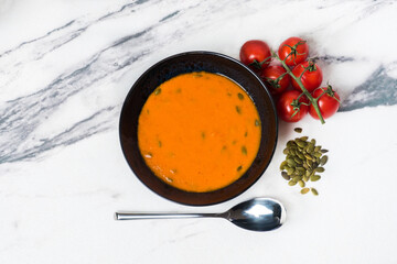 Vegetarian vegetable soup. Tomato and pumpkin soup with pumpkin seeds. A dark plate is on a white marble background. Nearby are cherry tomatoes. Diet. Proper nutrition. Light background