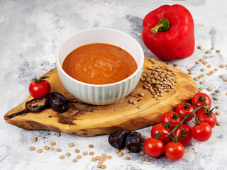 Vegetarian vegetable soup. Tomato soup with bell pepper and dates. A white deep plate stands on a wooden stand. Nearby are cherry tomatoes and lentils. Diet. Proper nutrition. Light background.