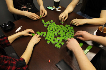 A team of four young people collect bones for playing mahjong on a dark, brown table. The back and side panels are blurred. Table traditional game of China, Japan, and other Asian countries.