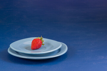 One small ripe red strawberry berry lies in a plate on a blue background