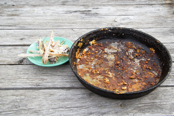 Black from the plaque very dirty frying pan stand on a wooden table. It was fried fish. Nearby is a saucer with bones from fish. Cooking at home