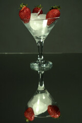 Tall martini glass with a pieces of ice and strawberry berries. Mirror background