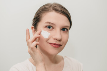 Happy young woman doing beauty treatment. The girl applies white cream on her face on the morning.