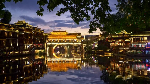 Fenghuang, China. Evening landscape with bright lights illumination. Bridge reflecting on Tuojiang river. People having a good time. Panning shot, UHD