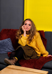 Portrait of girl in yellow oversize sweater, skirt and brown boots. Woman sitting on sofa. Yellow square on the background.