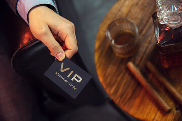 Man holds VIP member card. View from the top on the gentleman's hand that holds exclusive VIP membership card next to the wooden table with whisky in carafe and glass with cigars. - obrazy, fototapety, plakaty
