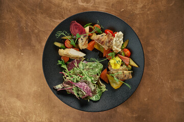 Appetizing warm salad with baked chicken fillet, bell pepper, tomatoes, asparagus and salad leaves in a black plate on a metal, copper background. Restaurant serving