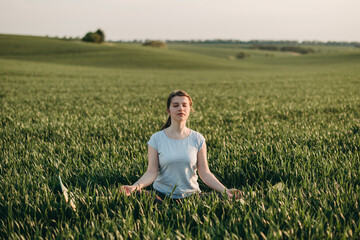 Young white woman practicing yoga in nature in a field of green grass in summer at sunset. Stretching, meditation, exercise, sun energy, meditation, breathing practice. 