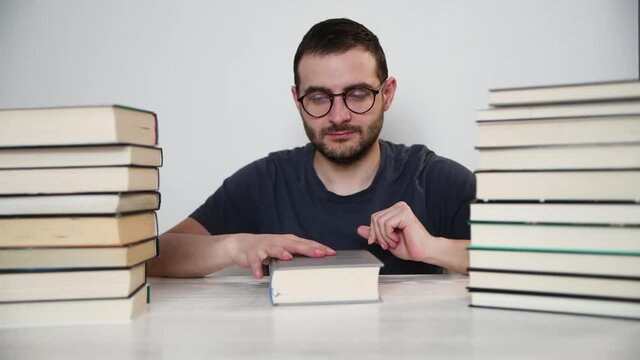 Young white European brunette man nerd in glasses teaches lessons, reads books, studied at university, prepares for exams, has many books at home in white backround table.