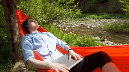 Cute freelancer woman weared in casual lying in hammock at the nature closing top of laptop relaxing and listening music in wireless headphones. IT specialist working outdoor