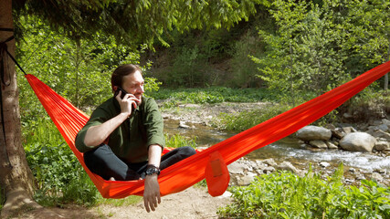 Man sitting in a hammock at the nature talking on smartphone. Scene of relaxing calm conversation outdoor