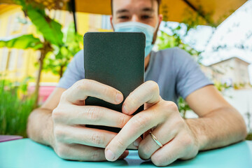 boy in a garden bar takeing a selfie with his face mask on to be protected from coronavirus - Teenager holding a smartphone sitting to do videocall outdoors in social distance from covid-19