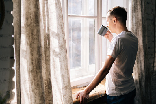 Pensive young man stands by the window on sunny day and takes sip of hot coffee or tea from cup, enjoying beautiful day.