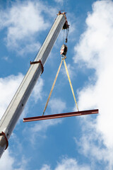 Construction crane with a hook and a metal profile hanging on ropes, low angle view with cloudy sunny blue sky © Aleksandra