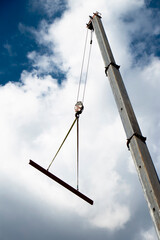 Construction crane with a hook and a metal profile hanging on ropes, low angle view with cloudy sunny blue sky - 354740555