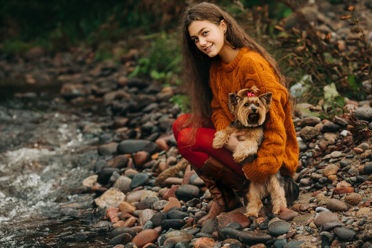 a girl in an orange sweater and red tights in the fresh air by a stream. walking with a dog Yorkshire Terrier - Powered by Adobe
