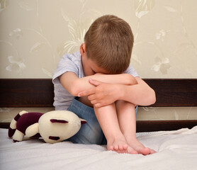 The child sits on the bed clasping his face and legs with his hands. Family violence. Child abuse.