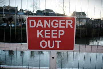 Generic Sign Reading 'Danger Keep Out' Seen on a Metal Fence of a Derelict Riverside Building with the Setting Sun in the Background