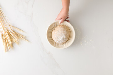 Top view of female hand holding a resting bread dough in a bread bowl in a marble table with wheat and flour with space for text