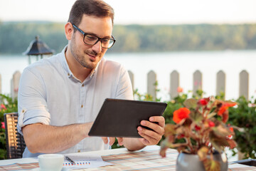 Happy young Caucasian freelancer drinking coffee in riverside cafe, browsing internet on a tablet while working on sales reports