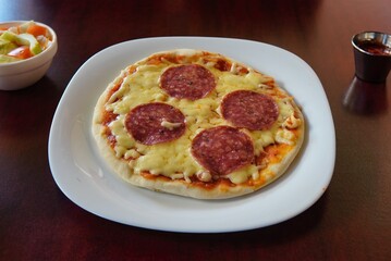 Closeup shot of a delicious mini pepperoni pizza with cheese and a salad on a wooden table