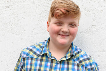 Plump red-haired boy is smiling. A boy in a plaid shirt stands on a wall background and smiles.
