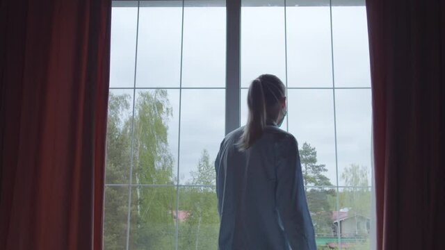 Girl in pajamas and a blue protective mask goes to the window and looks sadly at the street, sighs heavily. Girl in insulation complies quarantine