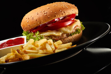 Big fresh burger with sauce and fries on black plate isolated on black background.  American classic burger menu.