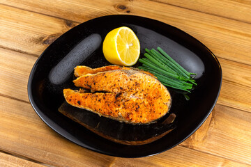 Grilled salmon steak with lemon slice and herbs in a black plate on a wooden background
