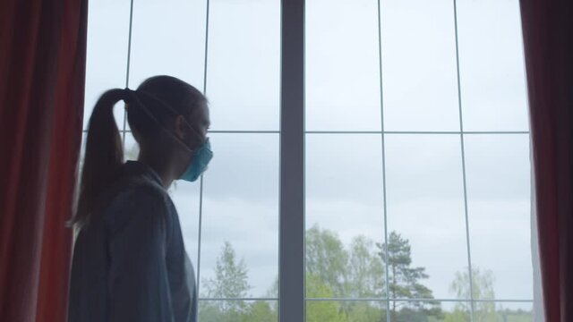 Girl in pajamas and a blue protective mask goes to window and looks sadly at the street, sighs heavily. Girl in insulation complies quarantine