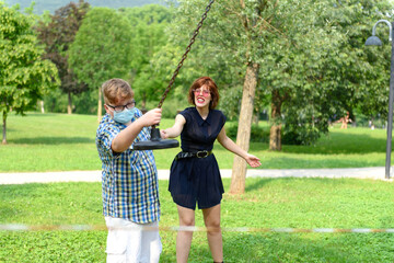 Obraz premium Boy wearing eyeglasses and surgical mask dressed in a plaid shirt plays with an adult girl in front of a red and white striped barrier tape around the playground. 