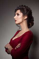 Fototapeta premium Beautiful woman with stylish haircut and big earrings looking down over shoulder, touching neck by hand. Brunette girl posing in dark studio, wearing red dress, with evening make up and long eyelashes