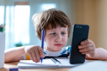 Naklejka premium Distance learning online education. Caucasian sad kid boy studying at home with smartphone and doing school homework. Thinking child siting at table with cell phone due coronavirus quarantine