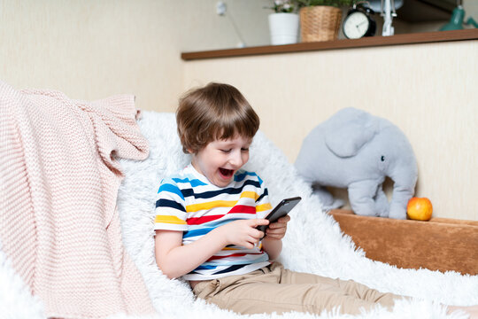 Happy little child boy playing online game, watching video on cellphone, sitting on couch entertaining in living room. Smiling small kid using funny mobile apps, enjoying free leisure time at home