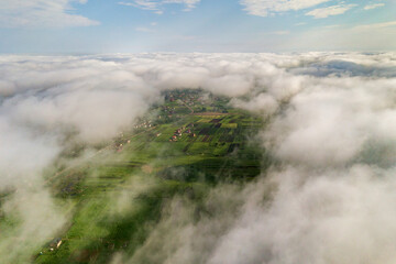Aerial view of white clouds above a town or village with rows of buildings and curvy streets between green fields in summer. Countryside landscape from above.