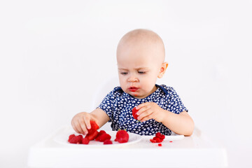 Happy baby girl sitting in high chair eating strawberry in a white kitchen. Healthy nutrition for kids. Bio fruits as first solid food for infant