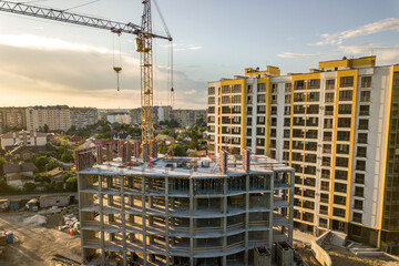 Apartment or office tall building under construction. Brick walls, glass windows, scaffolding and concrete support pillars. Tower crane on bright blue sky copy space background.