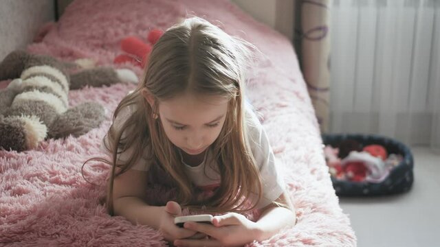 Girl plays the phone lying on the bed in her room.