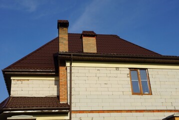 the attic of a white private brick house with a window under a brown tiled roof with chimneys against the blue sky on a sunny day