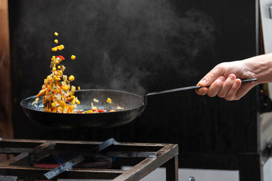 hand holding a frying pan, saut&eacute;ing fresh vegetables flying in the air