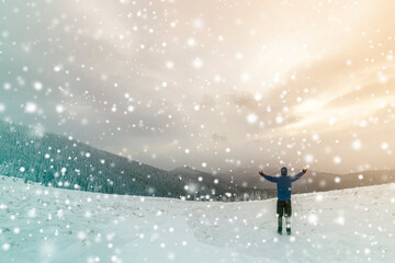 Back view of tourist hiker in warm clothing with backpack standing with raised arms on clearing covered with snow on spruce forest mountain and cloudy sky copy space background.