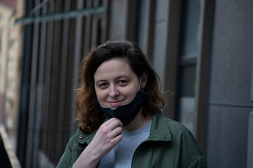 Smiling young woman removes the black protective mask on the street of a big city. Anti coronavirus, virus and flu concept. Back to normal life after pandemic. Quarantine is over. Urban evening scene.