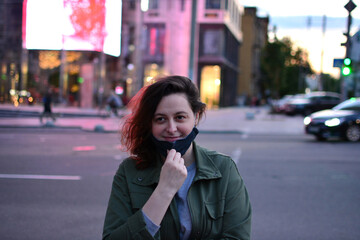 Fototapeta premium Smiling young woman removes the black protective mask on the street of a big city. Anti coronavirus, virus and flu concept. Back to normal life after pandemic. Quarantine is over. Urban evening scene.