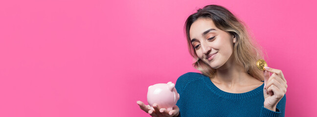 Girl puts in the piggy bank physical Bitcoin.Young girl over pink background holding piggy bank and rejoices gesturing. The concept of reliability of cash investments and insurance.