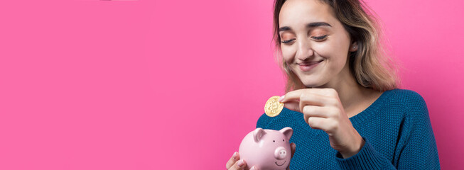 Girl puts in the piggy bank physical Bitcoin.Young girl over pink background holding piggy bank and rejoices gesturing. The concept of reliability of cash investments and insurance.