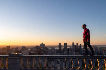 Fototapeta na wymiar Montreal, Canada - may 2020 : back view of a young man with a lumberjack shirt standing on a stone wall at the Kondiaronk belvedere and admiring the sunrise