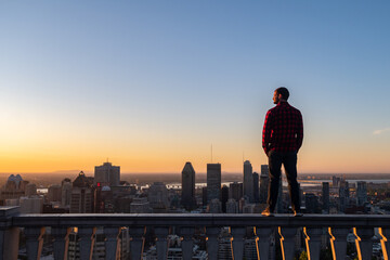 Montreal, Canada - may 2020 :  back view of a young man with a lumberjack shirt standing on a stone wall at the Kondiaronk belvedere and admiring the sunrise - obrazy, fototapety, plakaty