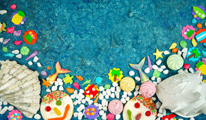 Summer beach theme flatlay with real and candy shells, pebbles, fish tails, summer party icons and cookie faces on a blue textured background with negative copy space.