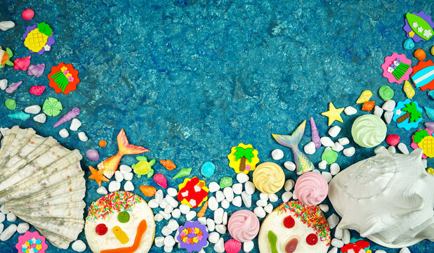 Summer beach theme flatlay with real and candy shells, pebbles, fish tails, summer party icons and cookie faces on a blue textured background with negative copy space.