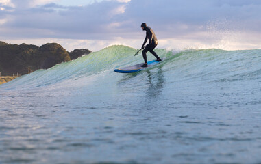 Naklejka premium Stand Up Paddle Boarding In Japan at Sunrise and Sunset a solo rider keeping fit & healthy on the Pacific Ocean in a black wetsuit, also catching some large waves, The ocean is blue with a nice sky.